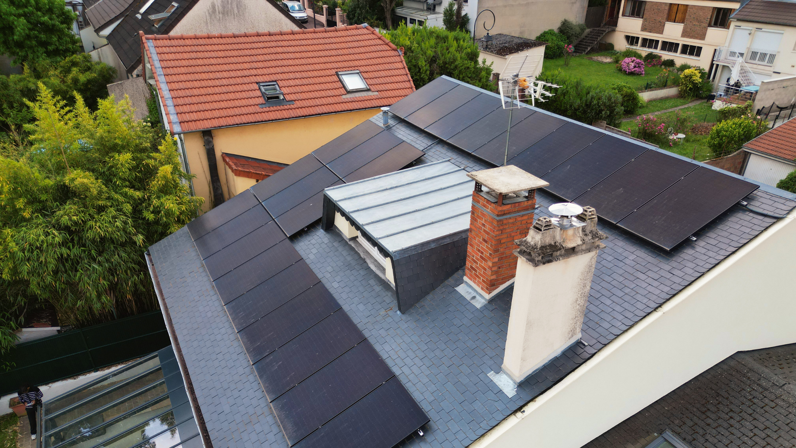 Toit d'une maison avec des panneaux solaires installés, entouré d'autres maisons et d'un immeuble en arrière-plan sous un ciel partiellement nuageux