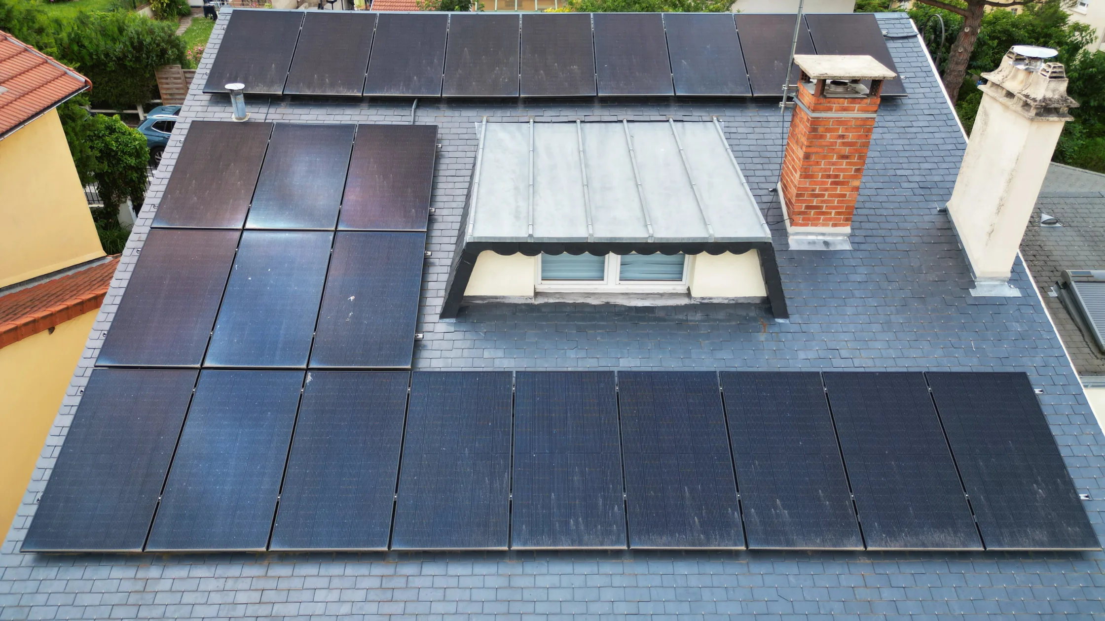 Vue rapprochée d'un toit de maison avec des panneaux solaires, une lucarne et une cheminée en briques.